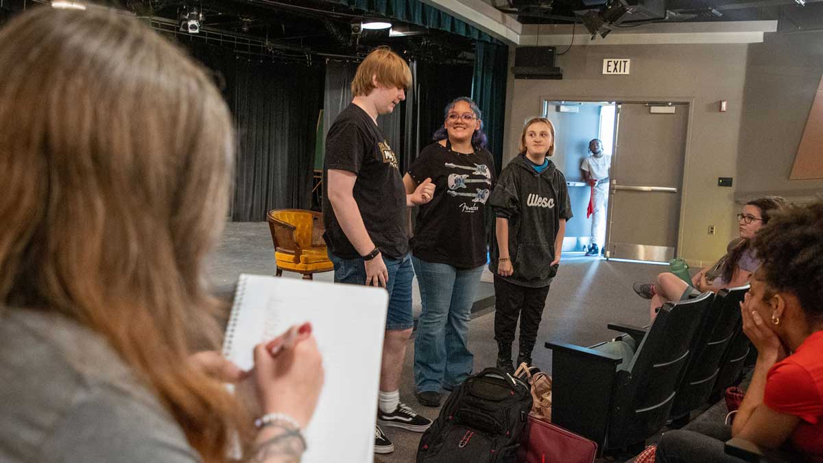 A group of students stand at the front of a small theater space while others watch from the audience, with one person sketching in the foreground.