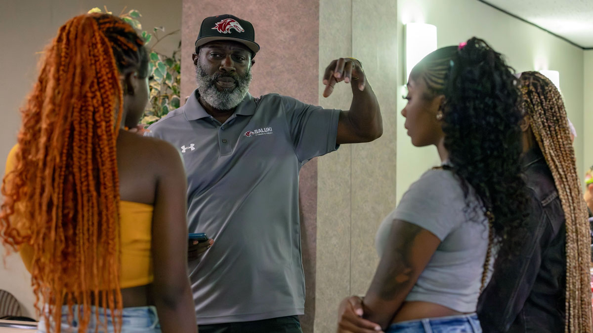 A staff member in an SIU polo speaks with three students in a hallway, using hand gestures as they listen.
