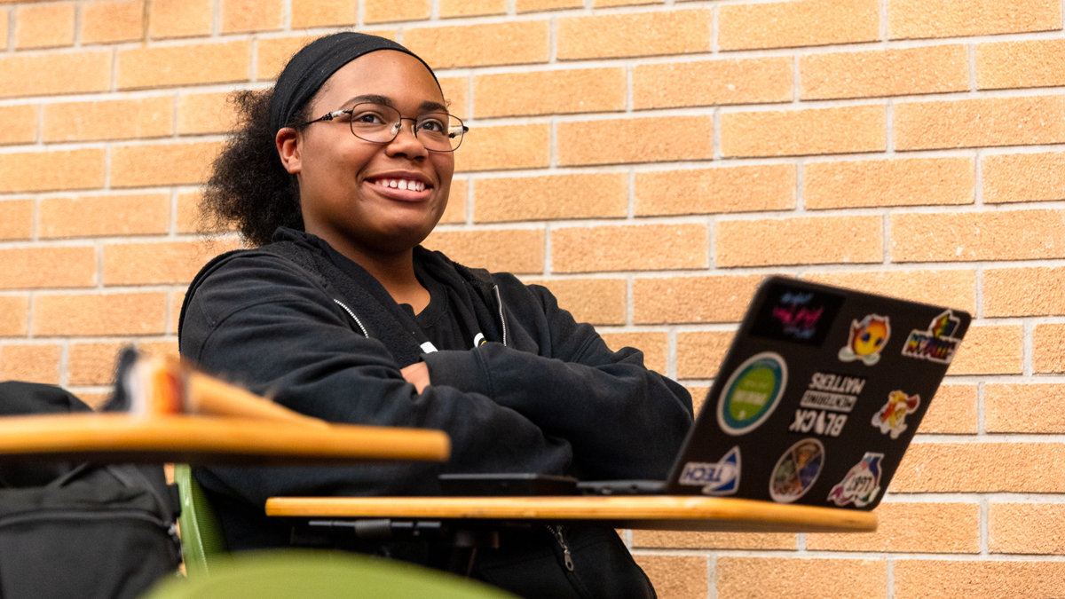 A student sits at a desk smiling with a sticker-covered laptop open in front of them.