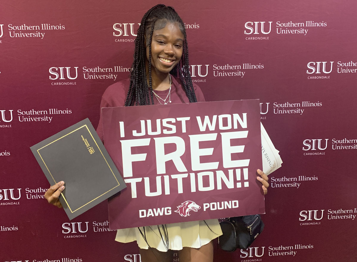 A student stands in front of an SIU backdrop holding a sign announcing they won free tuition.